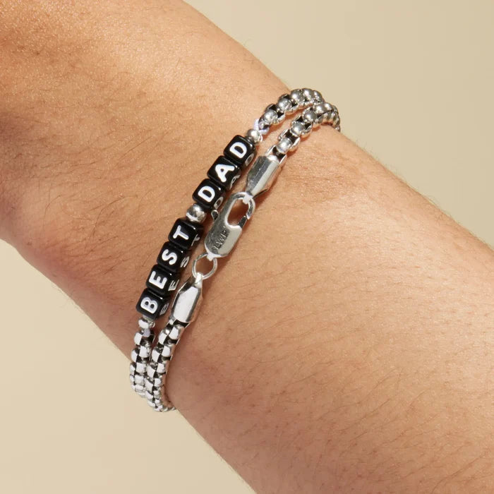 Silver box chain bracelet with black square letter beads spelling 'BEST DAD' worn on a wrist, stacked with a plain silver box chain bracelet. LWP-engraved silver lobster clasp visible in the foreground.