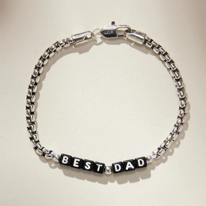 Silver box chain bracelet with black square letter beads spelling 'BEST DAD' at the center, laid flat on a cream background. Features an LWP-engraved silver lobster clasp closure.