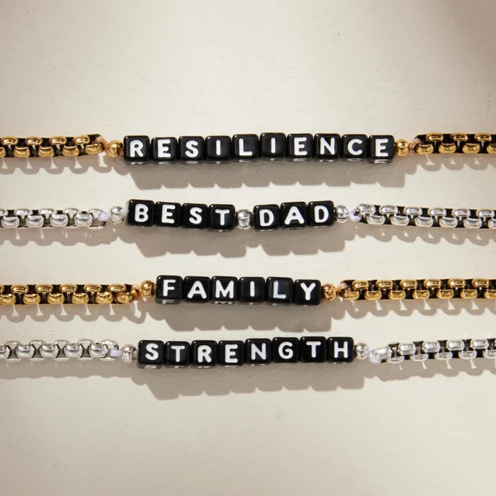 Four box chain bracelets laid horizontally on a cream background, each featuring black square letter beads spelling different words. From top to bottom: 'RESILIENCE' on a gold chain, 'BEST DAD' on a silver chain, 'FAMILY' on a gold chain, and 'STRENGTH' on a silver chain.