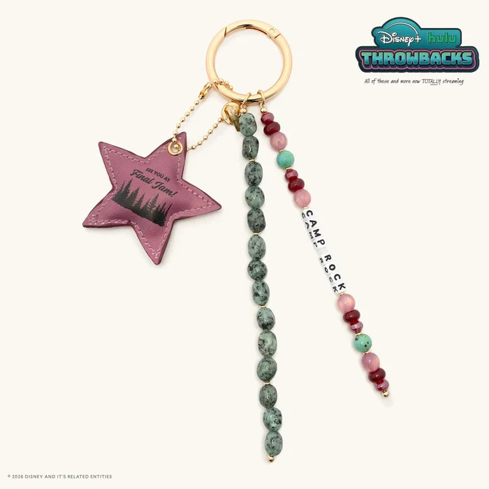 Camp Rock Disney+ Hulu Throwbacks bag charm with a gold ring, mauve faux leather star charm reading See You At Final Jam, green speckled stone beads, and Camp Rock white letter beads, flat lay on cream