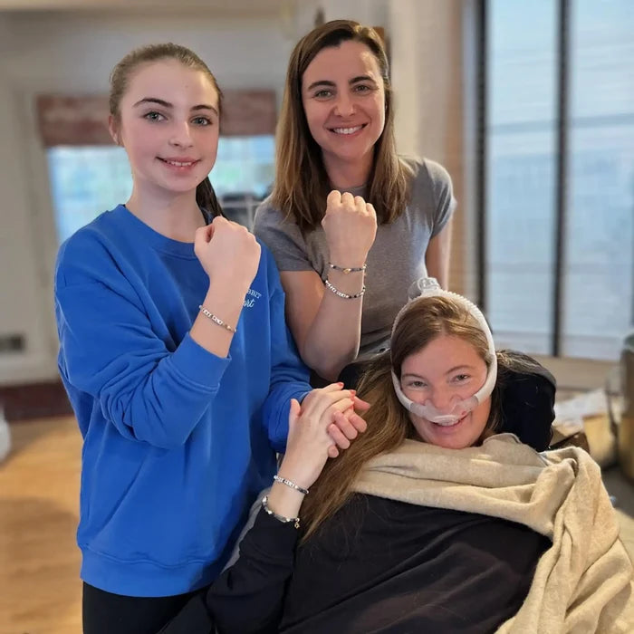 Three women smiling and showing off matching beaded bracelets, one seated and using a breathing tube.