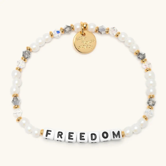 Freedom bracelet from the Project HEAL giveback collection, white faux pearl and clear and grey crystal beads with gold accents, black and white letter beads spelling FREEDOM, and a gold Little Words Project logo tag