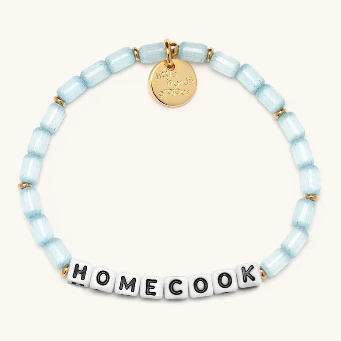 Homecook bracelet in Sunday Blues from the Little Words Project x Alex Snodgrass collaboration, with pale blue glass beads, gold spacers, and white letter beads spelling HOMECOOK, finished with a gold Little Words Project charm tag.