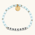 Homecook bracelet in Sunday Blues from the Little Words Project x Alex Snodgrass collaboration, with pale blue glass beads, gold spacers, and white letter beads spelling HOMECOOK, finished with a gold Little Words Project charm tag.