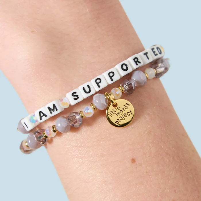 Two bracelets stacked on a wrist — one with white letter beads reading "I Am Supported" and one with grey, mauve, and iridescent crystal beads featuring a gold Little Words Project charm pendant.