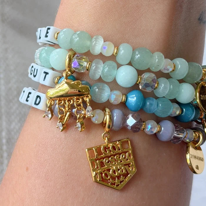 Multiple bracelets stacked on a wrist featuring letter beads (partially reading "Gut," "Ied/Ted," and "E"), mint green, teal, lavender, and iridescent beads, alongside various gold charms including a sneaker, a shield reading "I Got Dressed Today," and a small round pendant.