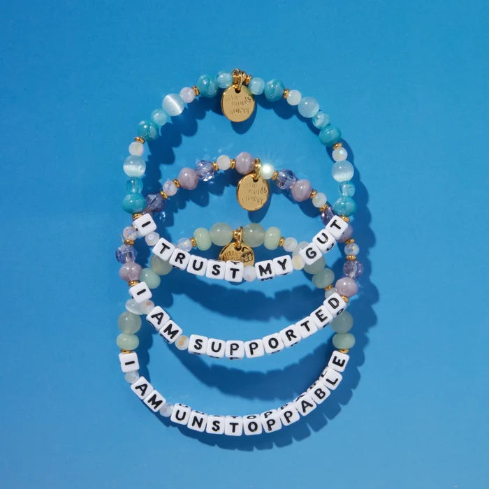 A stack of three stretch beaded bracelets arranged on a bright blue background. Each bracelet features white square letter beads spelling out affirmations: "I Trust My Gut," "I Am Supported," and "I Am Unstoppable." The beads between the letters are in soft pastel tones — mint green, lavender, blush pink, and translucent white — with small gold accent beads and round gold charm tags. The bracelets appear to be from the brand Little Words Project.