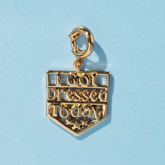 A gold shield-shaped charm reading "I Got Dressed Today" with decorative stars at the bottom, attached to a spring ring clasp, on a blue background.