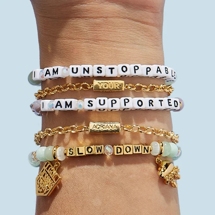 Four bracelets stacked on a wrist — white letter bead bracelets reading "I Am Unstoppable," "I Am Supported," and "Slow Down," interspersed with gold chain bracelets featuring personalized name bars reading "Your" and "Adriana," plus gold charms including the "I Got Dressed Today" shield and a decorative floral pendant.