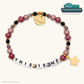 This Is Me Disney+ Hulu Throwbacks bracelet with burgundy, black, and dusty mauve pearl beads, white letter beads, and a gold star charm, flat lay on cream