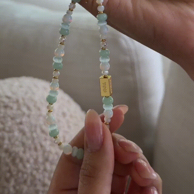 A gif of the Little Words Project beaded phone strap in soft blue, white, and gold tones with a spinning gold bar charm.