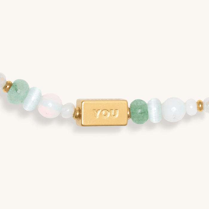 Close-up of a gold "Good" bar charm on a beaded strand with soft white, mint, and pink beads.